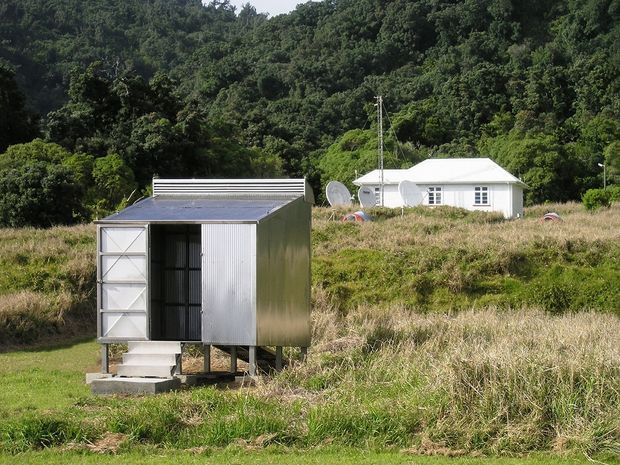 A small elevated corrugated-metal utility or storage structure stands in a grassy rural clearing, with a simple white gable-roofed house and dense wooded hillside in the background. The image is an exterior landscape view in daylight, showing a modest rural property and ancillary outbuilding.