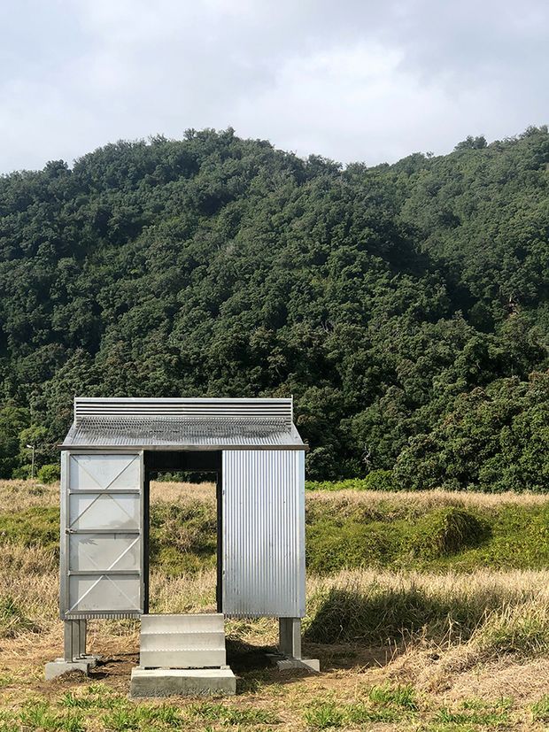 A small freestanding corrugated-metal shelter or utility structure stands in a grassy rural clearing, backed by dense native woodland and rolling hills. The structure has a simple rectangular form, a shallow corrugated roof with horizontal trim, a vertically ribbed metal side wall, and an open central doorway with a hinged metal panel door on the left. Concrete footings and a short concrete step support the elevated structure. The image is a straightforward outdoor architectural photograph in natural daylight.