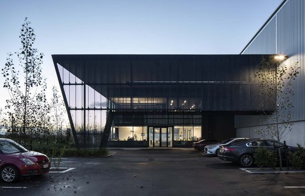 Exterior view of a contemporary multi-storey commercial building with a dramatic cantilevered dark metal canopy framing a glazed entrance. The facade combines extensive curtain-wall glazing with vertically ribbed metal cladding, and the building is approached via a paved parking area with landscaped planting.