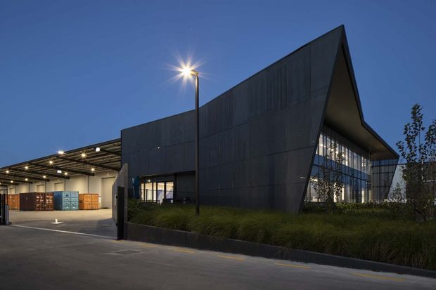 A dusk exterior view of a contemporary commercial or industrial building with a sharply angular, dark-clad facade and a dramatic sloping roofline. Floor-to-ceiling glazed sections reveal an illuminated interior, while a covered loading or service area with stacked shipping containers sits to the left. The building is set behind landscaped grasses and approached by a paved driveway, with exterior lighting creating a calm but dramatic architectural presentation.