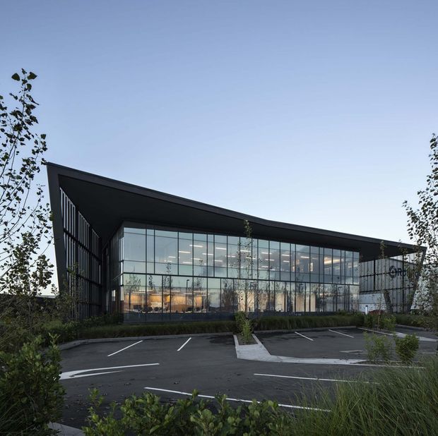 Exterior view of a contemporary commercial building with a dramatic asymmetrical, single-slope roof and dark structural framing. A large glazed curtain wall reveals illuminated office interiors, while landscaped planting and a marked car park occupy the foreground.