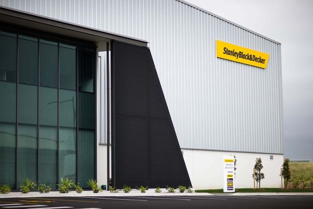 Exterior view of a large contemporary commercial or industrial building with a white vertically ribbed metal-clad facade, dark glazed entrance volume, and a prominent black angular architectural feature. A yellow StanleyBlack&Decker sign is mounted high on the facade, with landscaped planting and a paved access area in front.