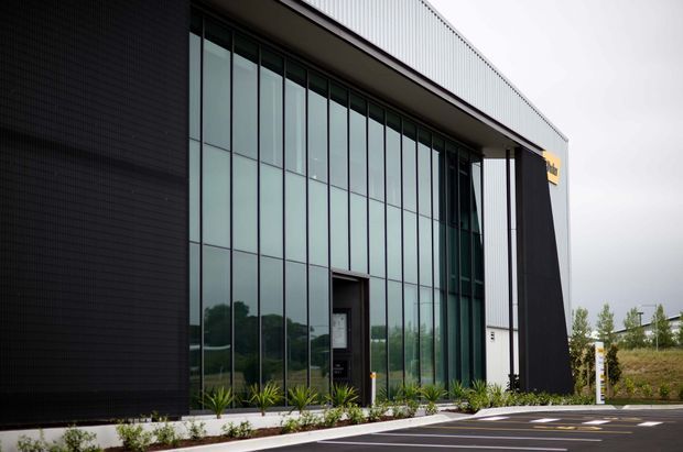 Exterior view of a contemporary commercial building with a tall glazed curtain-wall entrance, dark vertically textured cladding, a light ribbed metal upper facade, and a paved parking area with simple landscaping.