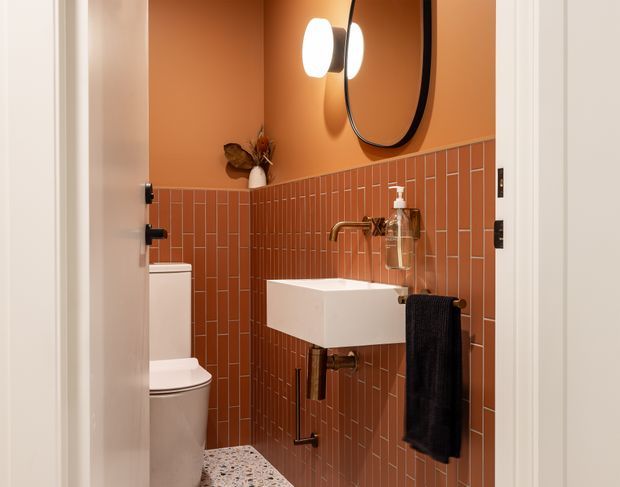 Compact contemporary powder room with terracotta-painted upper walls and matching vertical rectangular tiled wainscoting. A wall-mounted white basin with exposed bronze-toned plumbing sits beneath a rounded black-framed mirror and globe wall light. The room also includes a white toilet, black towel, small vase, and terrazzo-style floor.