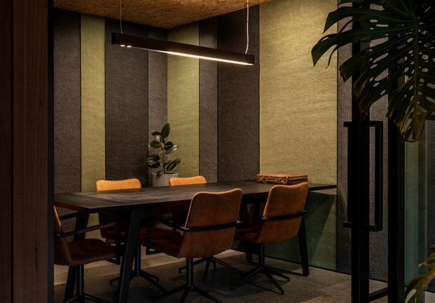 Moody contemporary dining area with a dark rectangular dining table and several brown upholstered chairs. The room features vertically paneled acoustic-looking wall surfaces in muted green, grey and brown tones, a textured warm ceiling, a slim linear pendant light, indoor plants, and black-framed glazed doors or partitions.