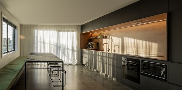 A spacious contemporary kitchen and dining area with a long dark island, slim black bar stools, built-in black cabinetry and appliances, and a warm timber backsplash with integrated open shelving. Floor-to-ceiling sheer curtains and large windows bring in abundant natural light, while a green upholstered bench runs along the dining side. The polished concrete floor and dark joinery create a refined, modern interior.