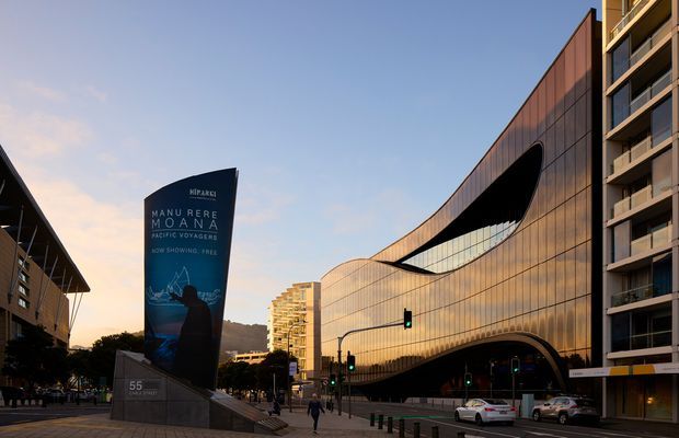 Street-level exterior view of a large contemporary performing arts and entertainment complex in an urban setting. The building has a sweeping curved facade clad in warm metallic panels, extensive dark glazing, and a dramatic oval-like recessed opening. A tall promotional sign and pedestrian plaza sit in the foreground, with a multi-storey apartment or commercial building adjoining the right side. The image is photographed in warm late-day light.