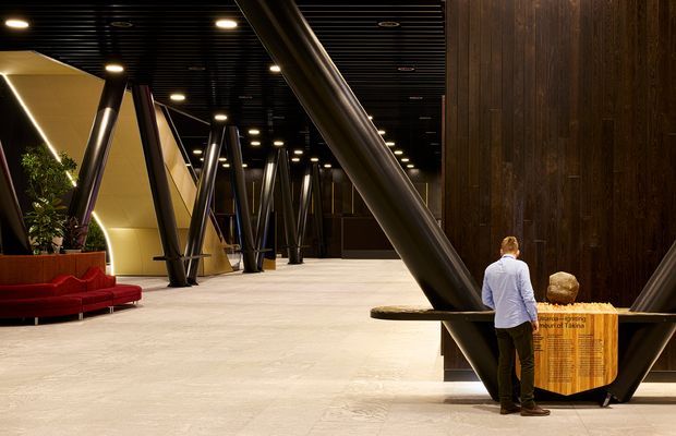 A dramatic contemporary interior lobby or commercial public space with a spacious pale stone or concrete floor, black slatted ceiling, integrated warm lighting, dark timber wall cladding, and prominent angled black structural columns. A red upholstered lounge sofa and indoor planting sit to the left, while a timber reception or information desk is positioned to the right. The high-contrast, warm-lit interior has a refined, monumental atmosphere.