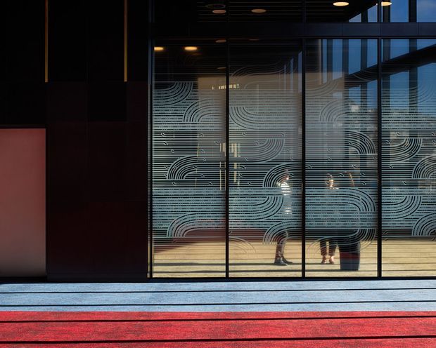 A dark, contemporary commercial entrance or lobby viewed from outside through full-height glazing. The glass is covered with a distinctive flowing linear patterned treatment, revealing silhouettes and reflections inside. Black metal framing, dark wall panels, warm recessed ceiling lights, and broad horizontal paving bands in grey and vivid red create a dramatic, graphic composition.