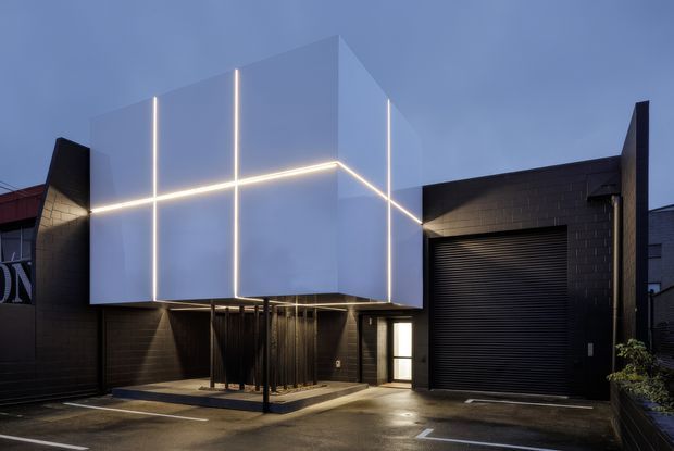 Contemporary dark-clad building exterior photographed at dusk, featuring a prominent cantilevered white geometric box volume outlined with integrated linear lighting. The ground level includes a covered entrance supported by slender columns, a glazed doorway, and a large black roller garage or loading door, with smooth paved forecourt.
