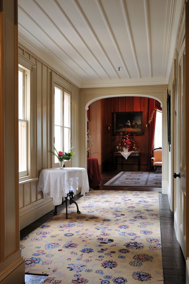 A refined traditional interior hallway or passage lined with tall windows, cream-painted wall panelling, ornate mouldings, and a white beadboard-style ceiling. A floral carpet runner extends toward an arched opening leading to a dark timber-panelled room with a framed artwork, console table, and floral arrangement. A small draped table and arrangement sit beside the windows.