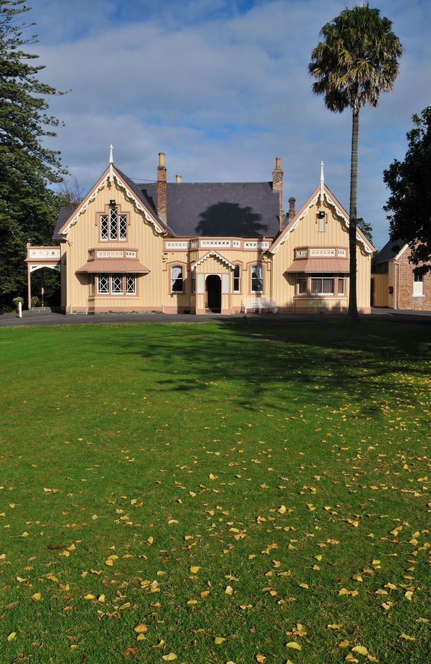 Front exterior view of an ornate historic timber-clad building with symmetrical gabled wings, decorative bargeboards, patterned window surrounds, brick chimneys, and a dark pitched roof. The building sits behind an expansive lawn with mature trees, including a tall palm, under a partly cloudy sky.