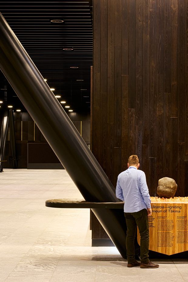 Interior view of a dark, sophisticated public exhibition or cultural space. A large diagonal cylindrical structural element crosses the room, set against a dramatic dark timber-panelled wall and black linear slatted ceiling with recessed lighting. Pale stone or concrete flooring contrasts with the dark surfaces. A visitor stands beside a sculptural display and interpretive signage, indicating a museum or gallery environment.