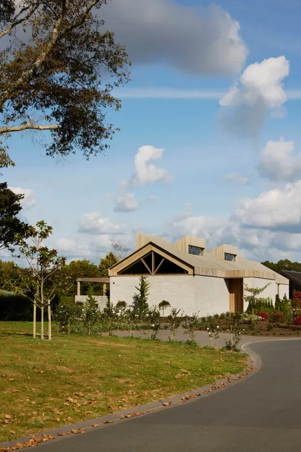 Exterior view of a contemporary low-rise building set within landscaped grounds. The structure has white brick walls, a pale metal gabled roof with several raised roof forms or clerestory-like elements, dark timber framing in the front gable, and timber-framed glazing or doors. A curved paved driveway and established trees surround the building under a bright partly cloudy sky.