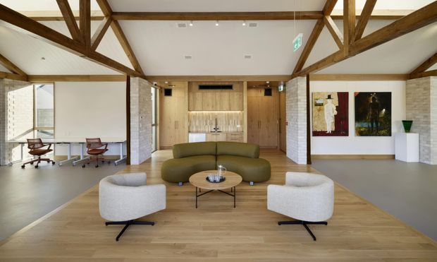 A bright, open-plan contemporary office lounge and reception area with exposed timber post-and-beam trusses, white painted brick walls, timber flooring, and a central curved olive-green sofa. Two white lounge chairs and a round coffee table form a seating arrangement in the foreground, while workstations are visible to the left and timber-lined service or kitchen joinery sits at the rear. Artwork and built-in display elements add visual interest.