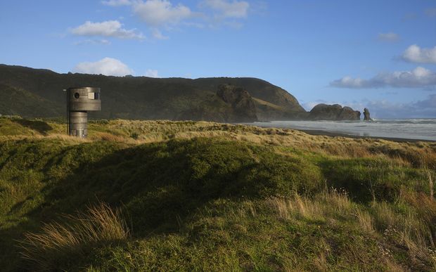 A coastal landscape with a cylindrical concrete lookout or observation structure set among sandy dunes and native grasses. Dark forested cliffs extend along the shoreline toward rocky sea stacks, with waves and a partly cloudy blue sky in the background.