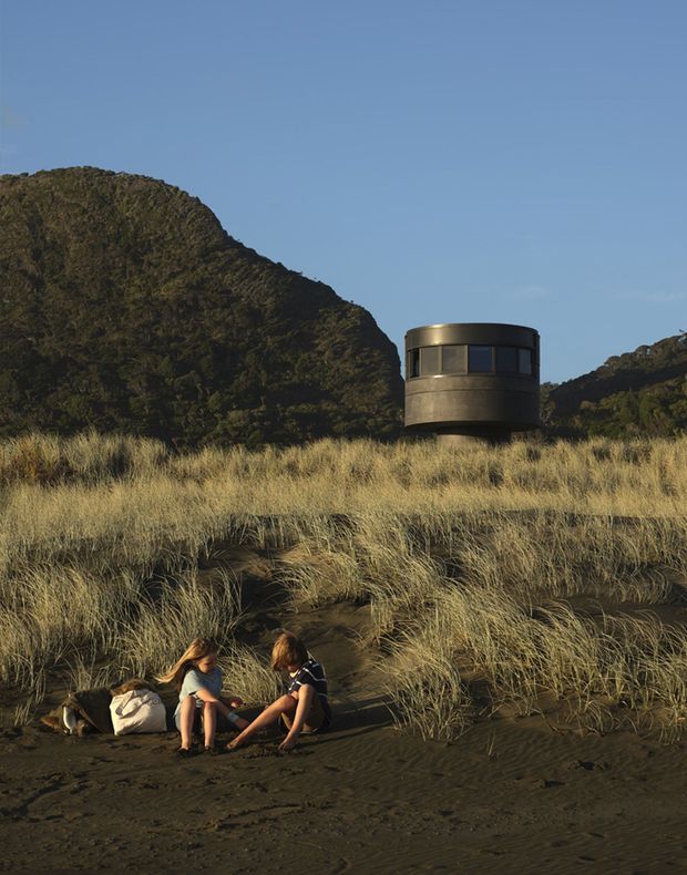 Exterior architectural photograph of a compact cylindrical, dark-toned building set among tall coastal grasses and black sand, with rugged forested hills behind it. Two people sit in the foreground, emphasizing the landscape setting and scale. The structure has a rounded form, a horizontal band of dark windows, and a smooth metallic-looking exterior.