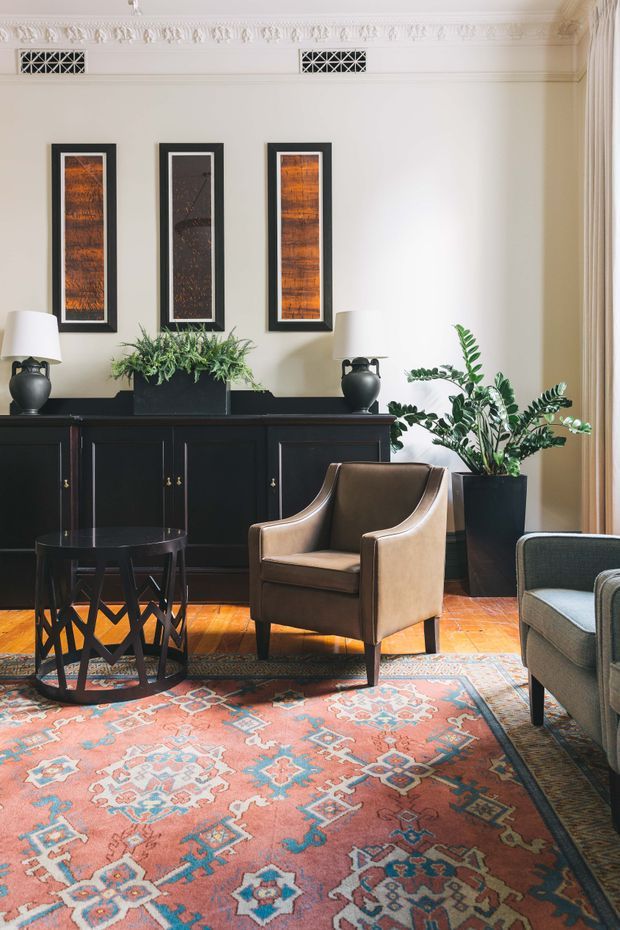 Elegant traditional sitting room with ornate ceiling cornices, cream walls, dark paneled sideboard, framed artwork, table lamps, indoor plants, leather armchair, upholstered seating, and a large patterned oriental-style rug over polished timber flooring.