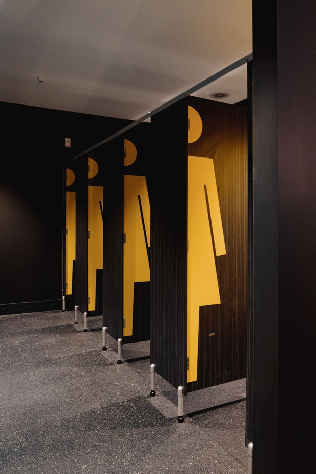 A dimly lit commercial restroom with multiple black and dark timber toilet cubicles. Each partition door features bold geometric yellow-orange graphics, creating a strong graphic and contemporary interior. The space has a speckled dark floor, black walls, a plain light ceiling with recessed fixtures, and exposed metal partition supports.