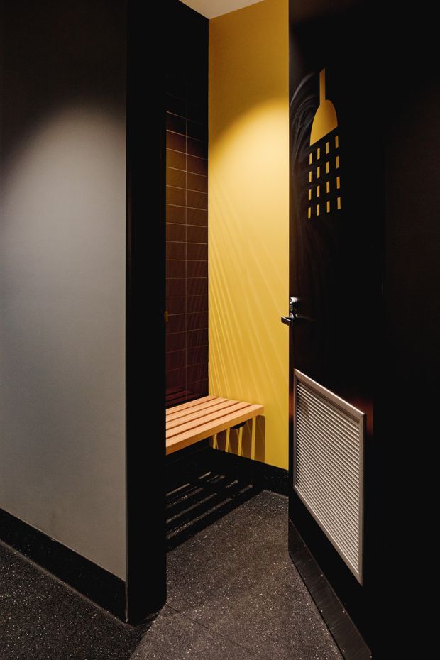 A dark, dramatically lit compact sauna or steam room entry. The space features black walls and door, a warm yellow textured wall, a slatted timber bench, dark tiled wall surfaces, and a metal ventilation grille on the open door. The atmosphere is moody and highly contrasted, with strong directional sunlight casting shadows across the floor and wall.