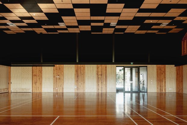 A large, empty indoor sports or multipurpose hall with a polished timber court marked by white lines. The walls combine vertical timber panels with pale acoustic sections, while the upper walls and ceiling are painted black. The ceiling features a dramatic geometric grid of suspended timber acoustic panels with integrated lighting. Glazed entry doors are visible at the far end.
