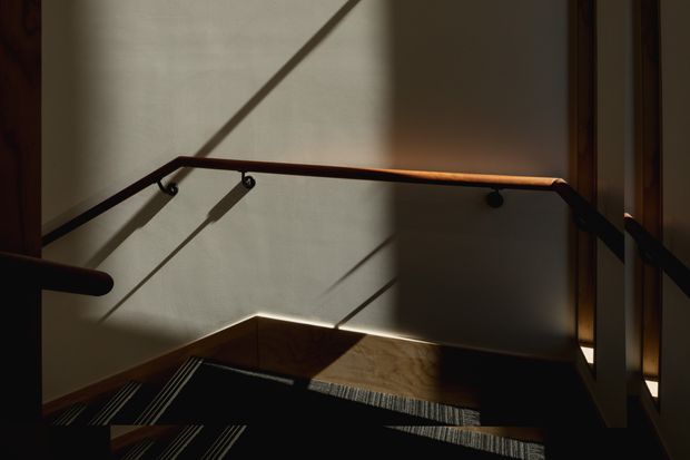 A dimly lit interior stairway or landing with dark carpeted steps, timber handrails, and slim metal supports. Strong sunlight casts long diagonal shadows across pale walls, while warm timber finishes and dark tones create a dramatic, refined atmosphere.