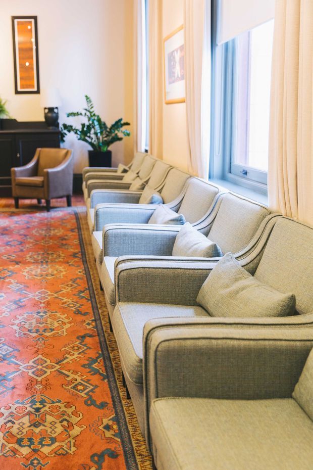 A warmly lit traditional sitting room or reception lounge with a long row of upholstered armchairs arranged beside tall windows. The room features cream walls and curtains, pale blue-grey seating with cushions, a vivid patterned orange and red carpet, framed artwork, dark cabinetry, and a potted plant. The composition emphasizes the seating area and abundant natural light.