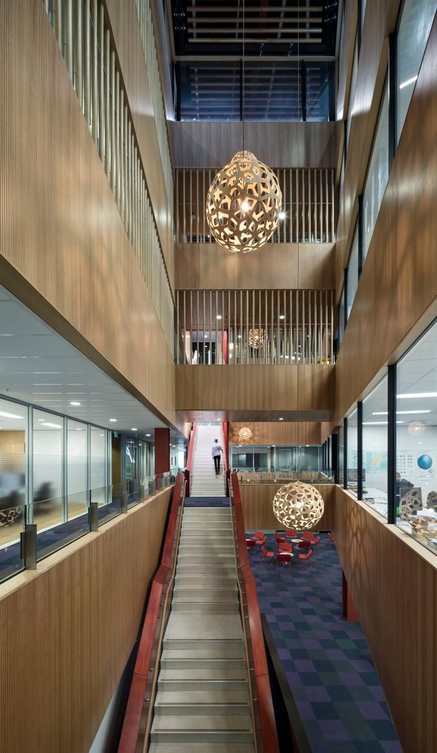 A dramatic multi-level commercial interior viewed along a central stairway. Warm wood wall panelling and vertical timber battens define the tall atrium, with glazed office or workspace fronts on both sides. Multiple suspended spherical perforated pendant lights hang through the void, while a patterned carpeted seating area is visible below. The space has a high glazed ceiling and strong vertical circulation axis.