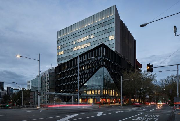 A dusk exterior photograph of a contemporary multi-storey commercial building on a prominent urban street corner. The building features a rectilinear glazed upper volume, a dramatic dark projecting lower facade with dense vertical fins, and a curved glazed entrance element. Traffic lanes, streetlights, signals, and surrounding city buildings reinforce the metropolitan setting.