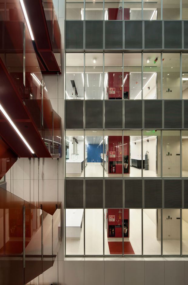 Vertical interior view of a modern multi-level commercial or institutional atrium. Repeated glass-fronted corridors and rooms are arranged across several storeys, with dark metal mullions, white illuminated interiors, and bold red structural or wall elements along the left side and within the central circulation zones. The space has a clean, highly geometric, contemporary architectural character.