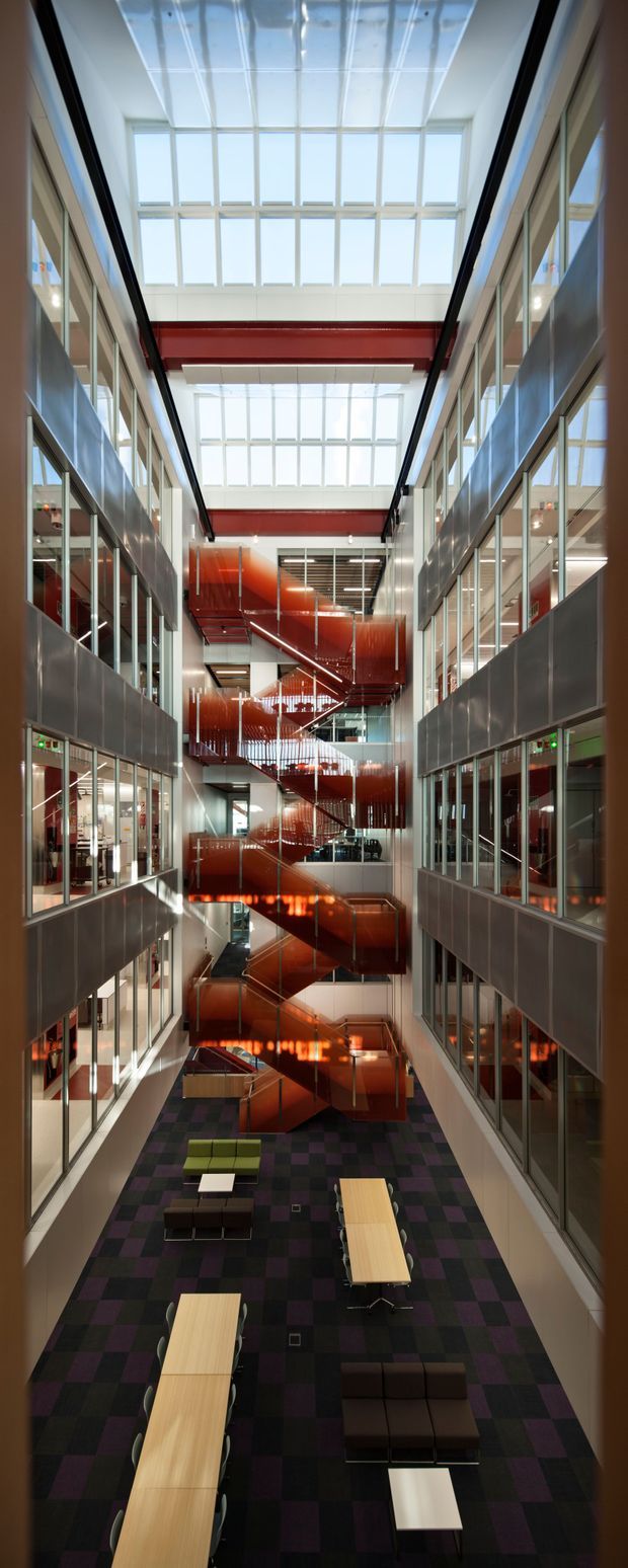 Tall interior atrium of a contemporary commercial or institutional building, viewed down through multiple levels. Glazed office or classroom fronts line both sides, while a striking red/orange multi-level stair and circulation structure occupies the centre. Large roof skylights and clerestory glazing flood the space with daylight. The ground level contains long light-wood communal tables, lounge seating, and a dark purple-black patterned carpet.