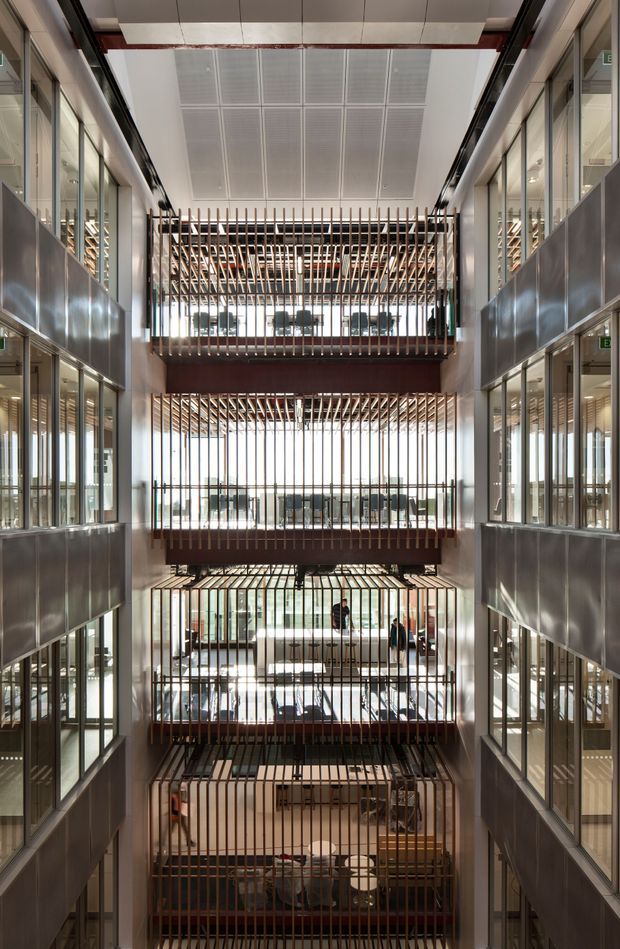 A tall, symmetrical interior atrium in a multi-storey commercial building, viewed across several levels. Floor-to-ceiling glazing lines both sides, while projecting walkways or balconies are screened by repeated vertical timber-toned slats. The central void is brightly daylit from a glazed roof or skylight, with exposed structural and services elements, metal panels, and office spaces visible on the surrounding floors. The architecture has a refined contemporary commercial character with strong vertical rhythm and layered transparency.