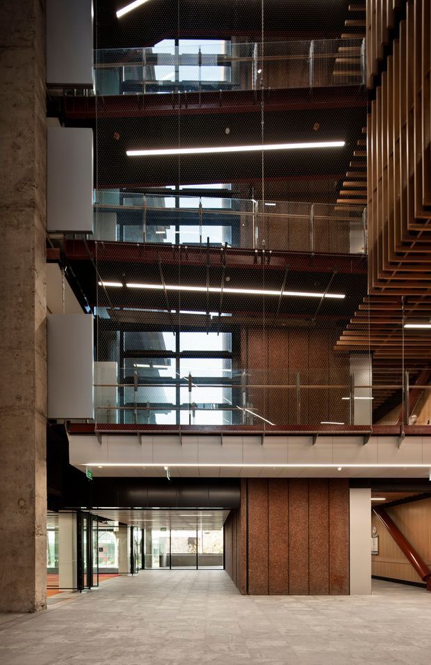 Interior view of a multi-level contemporary commercial atrium or circulation space. Several open mezzanine levels are stacked vertically, enclosed by transparent wire-mesh and glass balustrades, with exposed concrete columns, dark ceilings, linear strip lighting, reddish-brown metal or perforated wall surfaces, and prominent timber slat detailing on the right. The ground level opens toward glazed entry doors and shared circulation areas.