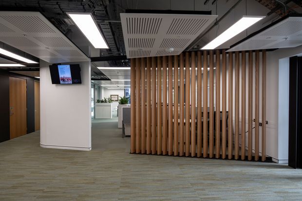 A contemporary commercial office interior with an open reception or circulation area, grey-green carpet flooring, white walls, exposed dark ceiling services, linear LED lighting, acoustic ceiling panels, and a prominent timber vertical-slat screen enclosing a workspace or meeting area. A digital display is mounted on a curved white column, with additional office workstations visible beyond.