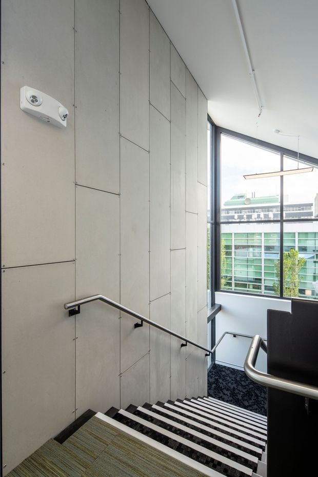 A contemporary interior stairway descending beside a tall concrete-panel wall. The stairs have dark treads with light horizontal nosings, black metal handrails, and a patterned dark carpeted landing. Large floor-to-ceiling windows provide abundant daylight and views of a neighbouring modern building. A linear pendant light and exposed ceiling conduit reinforce the modern commercial character.
