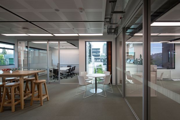 A bright contemporary open-plan commercial office interior with carpeted flooring, extensive glass-partitioned meeting rooms, open workstations, and breakout dining areas. The space features exposed ceiling services, linear acoustic ceiling panels, aluminium-framed glazing, timber communal tables, and white round tables with modern chairs. Urban buildings are visible through the windows.