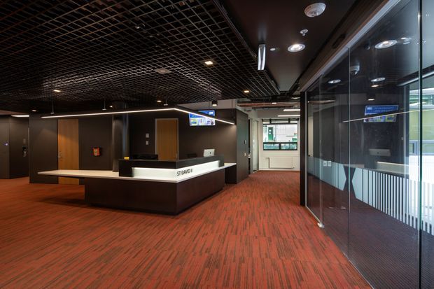 A dark, contemporary commercial reception lobby with a large angular reception desk, red patterned carpet, dark wood and charcoal wall finishes, extensive glass partitions, and a black exposed grid ceiling with recessed lighting. A small digital display and reception workstation sit behind the desk, while glazed doors and windows open toward a brighter exterior area.