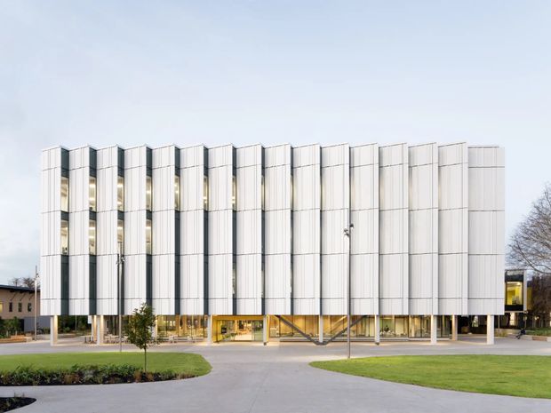 Exterior view of a contemporary multi-storey commercial building with a highly regular facade of vertical white metal panels and narrow glazed openings. The elevated upper volume is supported by slender columns, with a transparent glazed ground level and landscaped lawns and paved access in front.