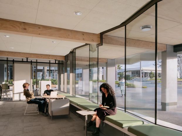 A bright contemporary commercial interior with a glazed perimeter and informal lounge or waiting areas. Floor-to-ceiling black-framed glass partitions and windows overlook a landscaped civic or campus setting. Green upholstered banquette seating, small tables, lounge chairs, and several people using laptops occupy the space. Exposed timber beams contrast with a pale tiled ceiling and carpeted floor.