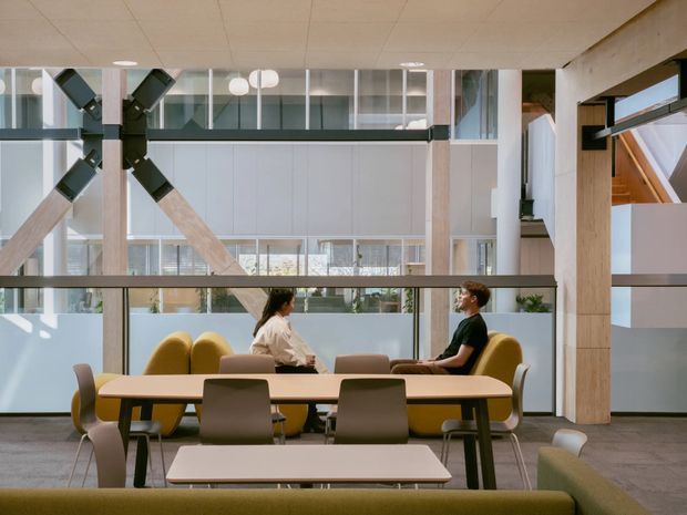 A bright contemporary communal interior with a large shared dining or meeting table, upholstered chairs, and lounge seating. Exposed timber and dark structural bracing frame a glazed, multi-level atrium with abundant internal daylight, planting, and people seated in conversation. The space appears to be a commercial or institutional social area rather than a residential room.