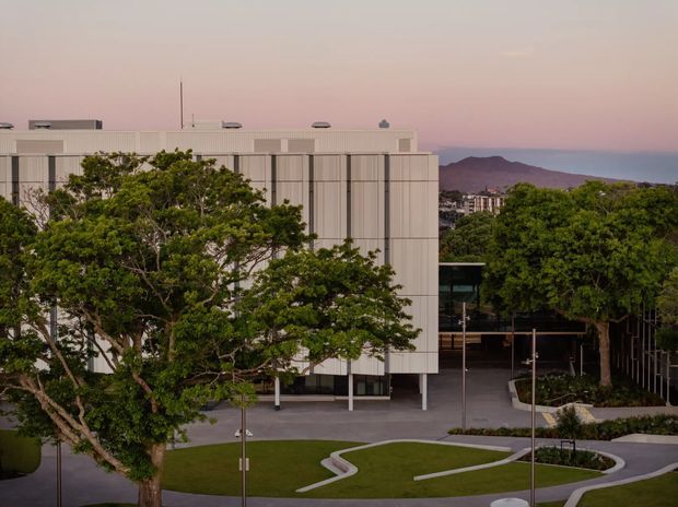 Exterior view of a contemporary institutional or commercial building with a pale vertically panelled facade, flat roof, extensive glazing, and a landscaped forecourt. Mature trees partially screen the building, while curving concrete paths and clipped lawn define the site. A coastal mountain ridge and urban buildings are visible in the distance under soft pink evening light.
