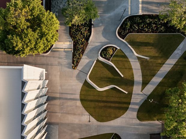 Overhead aerial view of a landscaped architectural site with curving concrete pathways, sculpted lawn areas, planted garden beds, mature trees, and a white sawtooth-roof building edge at the lower left. The composition emphasizes organic circulation patterns and geometric landscape planning.