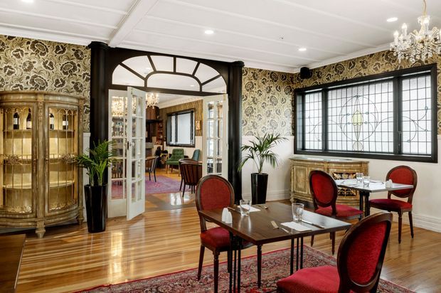 Elegant formal dining room with polished timber flooring, richly patterned wallpaper, ornate dark timber architraves, a large leaded-glass window, vintage-style dining furniture with red upholstered chairs, an antique display cabinet, a decorative sideboard, and a crystal chandelier. An adjoining sitting or living area is visible through the glazed arched doorway.