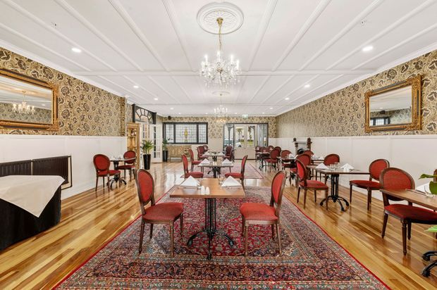 A spacious, ornate dining or hospitality interior with rows of small tables and red upholstered chairs arranged over richly patterned Persian-style rugs. The room features polished timber flooring, patterned wallpaper, white decorative wall panelling, large gilt-framed mirrors, a coffered and moulded ceiling, recessed lighting, and crystal chandeliers.