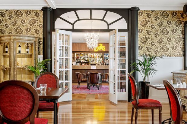 A refined restaurant or hotel dining room with polished timber flooring, ornate patterned wallpaper, dark architectural columns, white wainscoting, and red upholstered dining chairs. Glazed French doors open to a small bar area with a chandelier, backlit signage, and additional seating. A glass-fronted display cabinet and indoor palms add decorative detail.