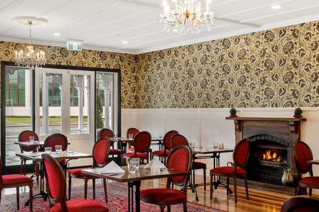 A refined traditional dining room or restaurant interior with numerous small tables and red upholstered chairs, ornate patterned wallpaper, white wainscoting, polished timber flooring, crystal chandeliers, and a central fireplace with a timber mantel. Glazed entrance doors open to an exterior courtyard or street beyond.