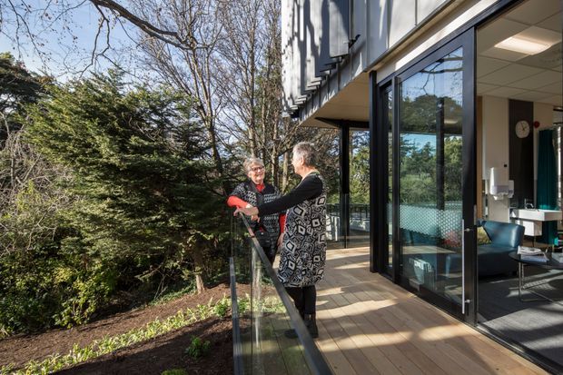 Photograph of a contemporary building viewed from an outdoor timber deck or balcony, with two people standing beside a frameless glass balustrade. Large black-framed glazed doors open into a bright interior lounge or office-like space, while mature trees and landscaped planting surround the building.