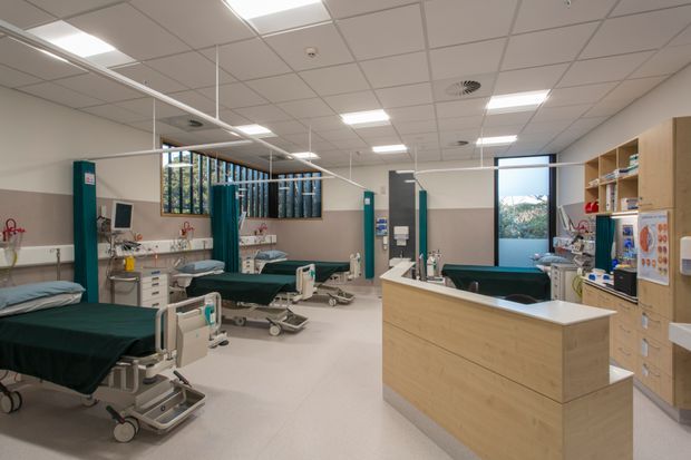 A bright, modern hospital treatment or recovery ward with multiple beds, teal privacy curtains, medical equipment, wall-mounted services, and a light timber reception or nurse station. The space has a suspended white acoustic tile ceiling with recessed fluorescent lighting, pale flooring, and built-in storage and work surfaces.