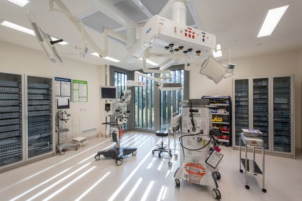 Bright, high-tech clinical procedure room with ceiling-mounted medical equipment, patient monitoring machines, mobile carts, stools, storage cabinets, and large glazed doors providing strong daylight. The space has clean white finishes and a highly functional hospital or healthcare fit-out.