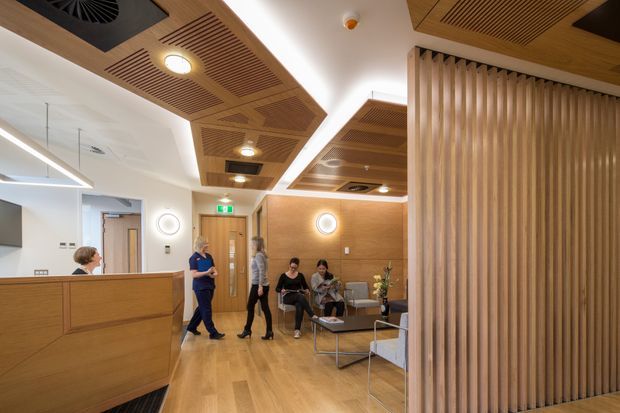 A warm, contemporary commercial reception and waiting area, likely in a healthcare or office setting. The space features extensive timber-look wall panelling, a timber reception desk, vertical timber screening, coordinated slatted timber ceiling panels with integrated lighting and ventilation, pale timber flooring, and a small seating area. People are present near the reception and waiting zone, with a corridor and illuminated exit sign in the background.